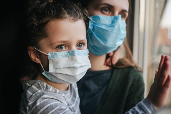 Unhappy young mother embracing upset little curly daughter with virus mask, sitting on windowsill at home, consoling sad preschool girl. Concept of coronavirus or COVID-19 pandemic disease symptoms